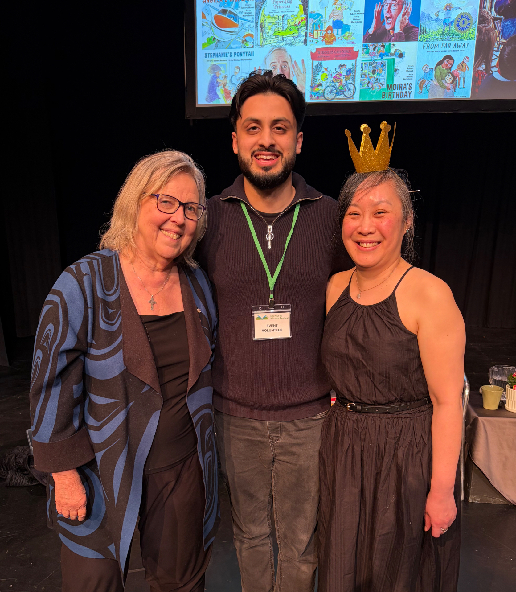 Mustafa Zuberi as an event volunteer at the Eden Mills Writers' Festival, standing with two attendees in front of a screen showing Robert Munsch book covers
